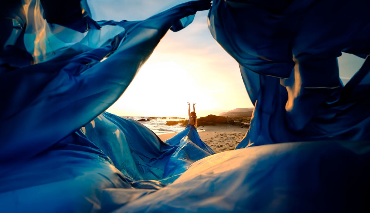 Services A woman poses elegantly in a blue dress on a beach at sunset, framed by flowing fabric.