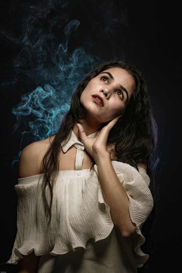 About Beautiful serene brunette in stylish white outfit with bare shoulders touching face tenderly and looking up while standing in dark smoky studio