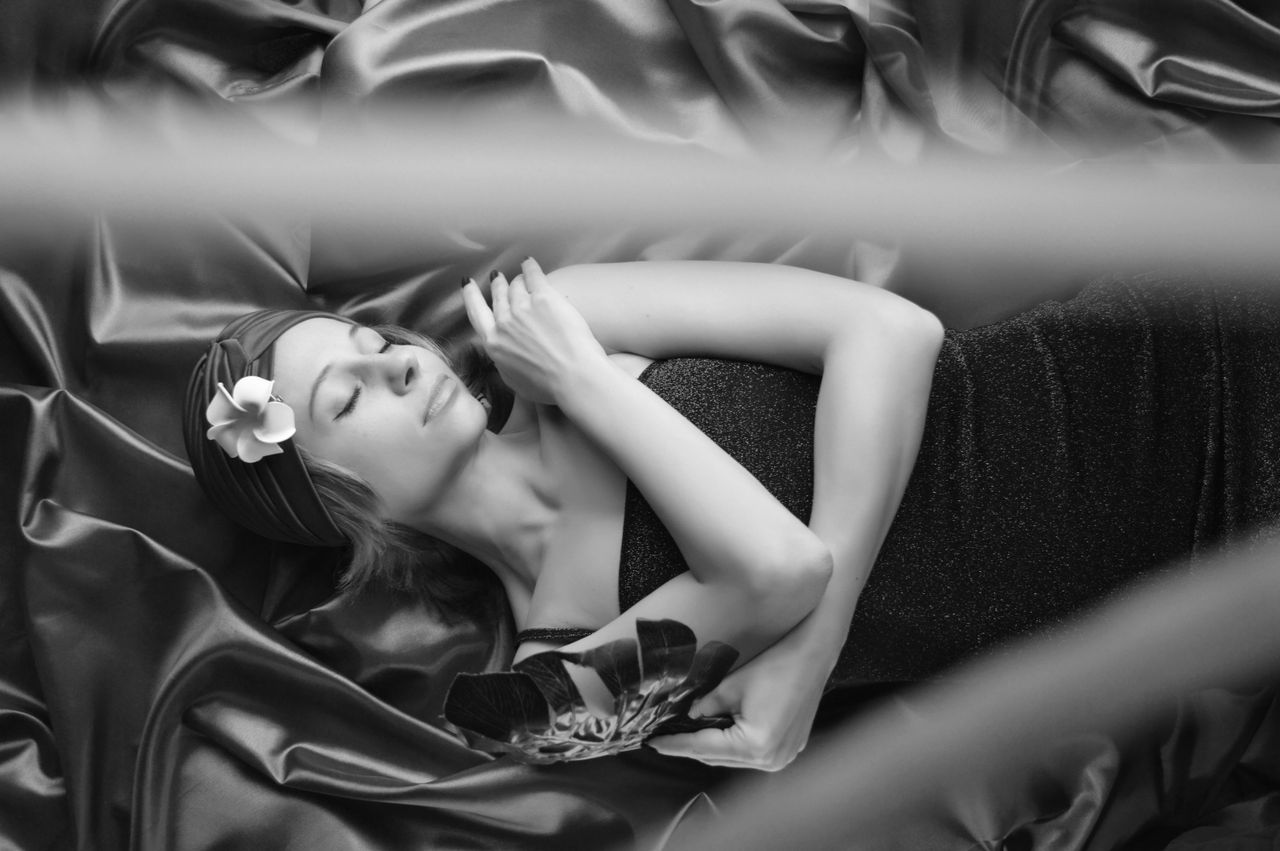 Black and white top view of young mindful female with blossoming Plumeria touching shoulder while lying on smooth creased textile